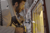 A BWXT Medical employee, wearing a white coat and safety glasses, using robotic manipulators in front of a hot cell window.